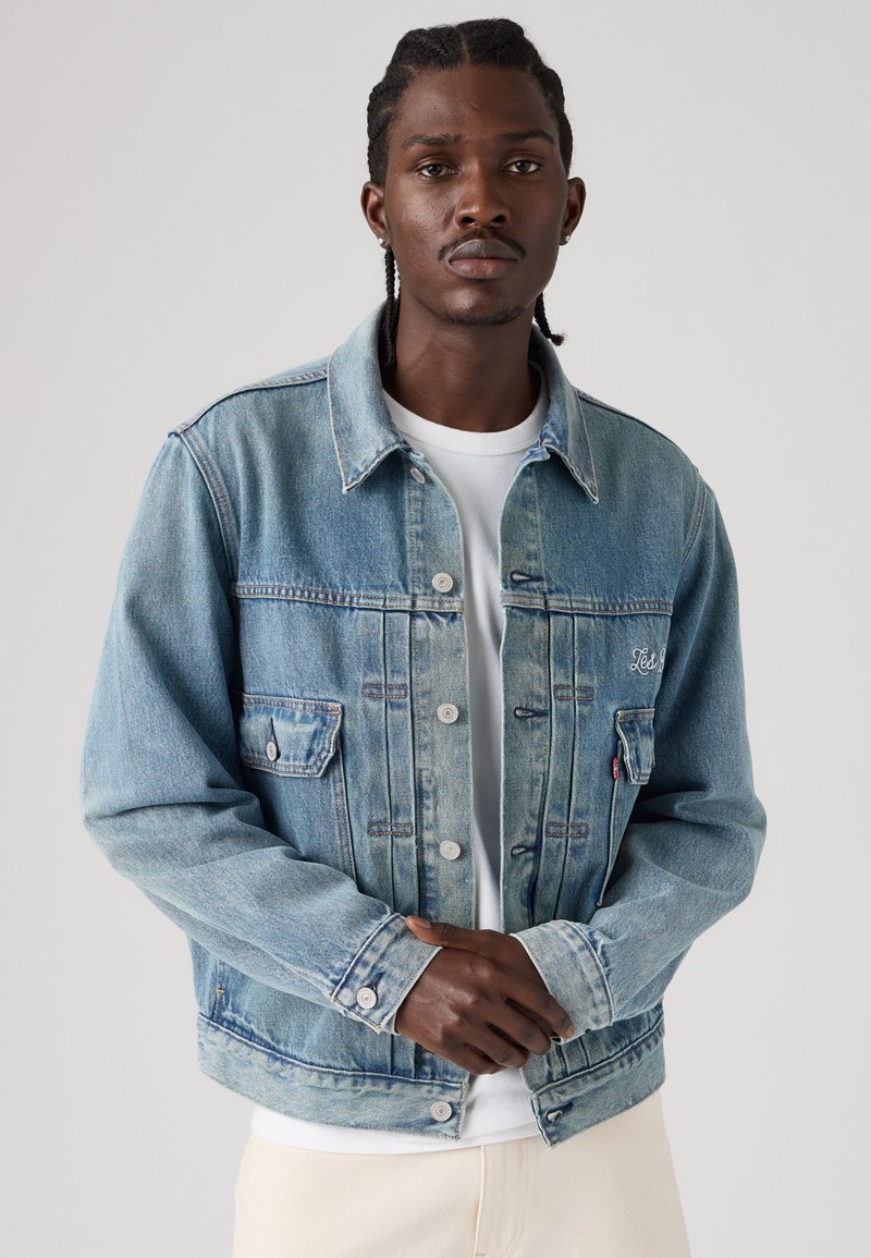 Giovane uomo con capelli intrecciati che indossa una giacca di jeans azzurro chiaro sopra una camicia bianca, in piedi davanti a uno sfondo grigio semplice.