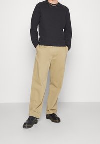 Zwarte sweatshirt gecombineerd met beige chino broeken. Model draagt zwarte veterschoenen. De setting is neutraal om de kleding te benadrukken.
