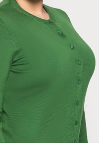 Groene gebreide cardigan met knoopsluiting, voorzien van een ronde hals en geribbelde textuur. Close-up toont materiaal en kleur.
