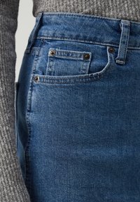 Denim-nilkkapituiset farkut, joissa korkea vyötärö ja keskiharmaa sininen väri. Farkuissa on viisi taskua, kuparinsävyiset metalliosat, ja osittain näkyvissä on teksturoitu harmaa neulepusero.