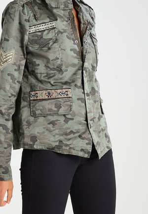 Donna che indossa giacca mimetica verde con toppe ricamate e pantaloni neri su sfondo semplice.