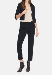 Black cropped jacket over a beige tank top, paired with high-waisted black trousers and brown ankle boots with a pointed toe and zipper detail.