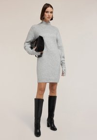 Abito maglione grigio oversize con collo alto e maniche lunghe, abbinato a stivali neri lucidi alti fino al ginocchio con tacco e una borsa nera a spalla.