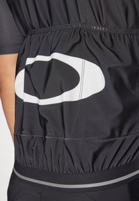 Schwarzes Radtrikot mit elastischem Bund, ausgestattet mit einem großen weißen Logo und strukturiertem Stoff. Die Nähte sind zur Langlebigkeit verstärkt.
