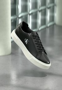 Μαύρο χαμηλό αθλητικό παπούτσι με λευκή σόλα και λογότυπο "CK Calvin Klein" στην πλευρά, σε φόν γυάλινης τοιχοποιίας.