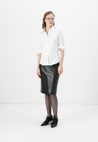 Camicia bianca con maniche arrotolate, abbinata a una gonna a matita in pelle nera. Si indossano scarpe nere con lacci e collant trasparenti.