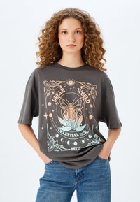 Camiseta gris de gran tamaño con un diseño gráfico colorido de una chica y elementos celestiales, con un escote redondo y mangas cortas.
