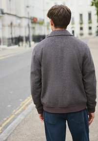 Brown checkered bomber jacket with a ribbed collar and cuffs, paired with dark blue straight-leg jeans. Street setting in the background.