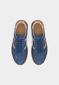 Ζευγάρι μπλε σουέντ αθλητικά παπούτσια με μπεζ ρίγες και καφέ σόλες, σε θέα από πάνω σε λευκό φόντο.