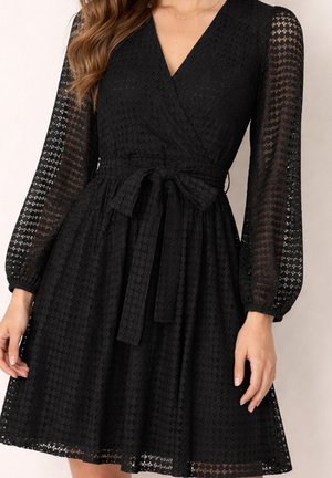 Femme portant une robe noire texturée avec manches longues transparentes, encolure en V et un nœud noué à la taille, montrant le haut du corps et les mains.