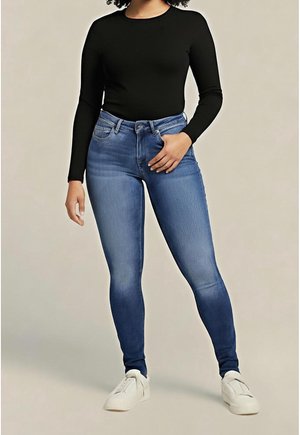 Vrouw draagt een zwart shirt met lange mouwen, blauwe skinny jeans en witte sneakers, staand met één hand in haar zak tegen een effen achtergrond.