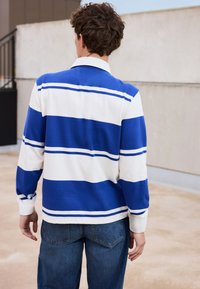 Langärmliges Polohemd in Blau- und Weißstreifen, mit einem Kragen und Knopfdetails am Hals, kombiniert mit blauen Jeans.