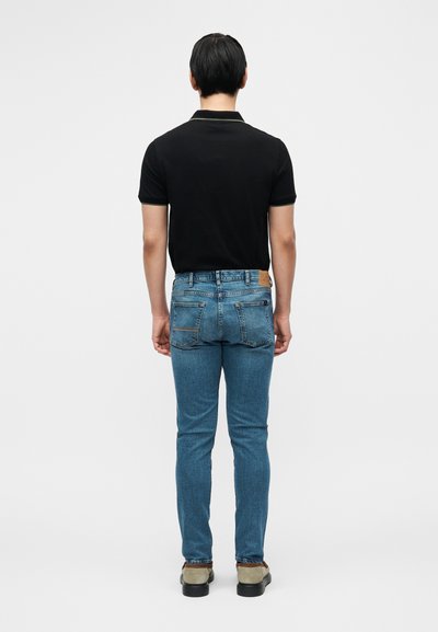 Μαύρο πόλο πουκάμισο, μπλε τζιν στενής γραμμής με πίσω τσέπες, δερμάτινο έμπλαστρο στη μέση και καφέ παπούτσια — προβολή από πίσω.