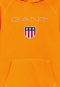 Oranžová mikina s kapucí, přední kapsou, vyšitým logem "GANT" a nášivkou s třemi hvězdami a červenými, bílými a modrými pruhy.