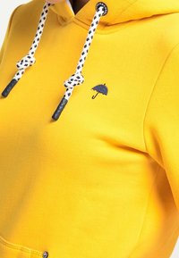 Sweat à capuche jaune avec un petit emblème de parapluie bleu marine, des cordons de serrage à carreaux noirs et blancs contrastants et une texture douce.