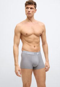 Homme torse nu portant un boxer gris Lacoste, debout et regardant légèrement vers sa gauche sur un fond clair uni.