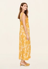Gelbes Blumenkleid im Maxiformat mit breiten Trägern, fließendem Design und strukturiertem Stoff. Kombiniert mit goldenen, spitz zulaufenden Flats.