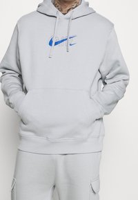 Světle šedá mikina s kapucí vyrobená z měkkého materiálu, s centrálním modrým logem Nike Air, klokaní kapsou a nastavitelnou kapucí se šňůrkou.
