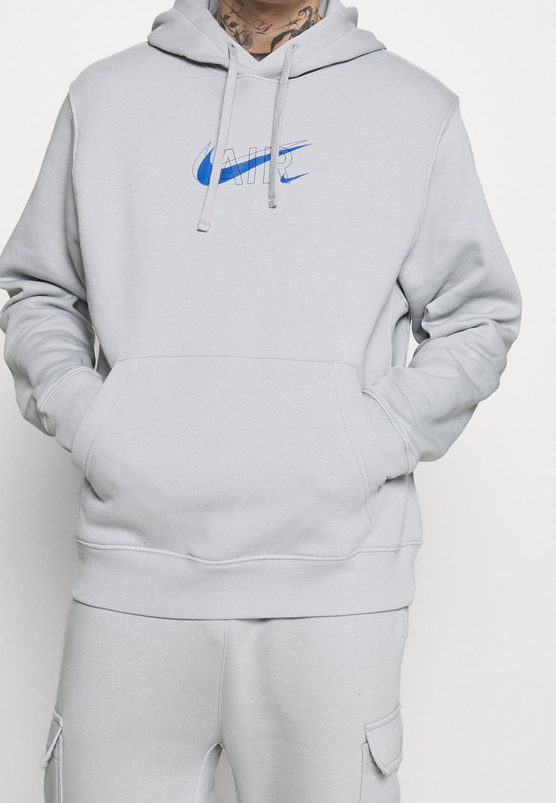 Světle šedá mikina s kapucí vyrobená z měkkého materiálu, s centrálním modrým logem Nike Air, klokaní kapsou a nastavitelnou kapucí se šňůrkou.