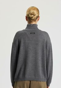 Maglione grigio lavorato a maglia con colletto alto e spalle scese. Presenta un piccolo etichetta del marchio sul retro, tessuto testurizzato e orlo a coste.