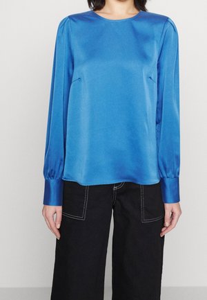 Persoon die een glanzende blauwe blouse met lange mouwen en gerimpelde manchetten draagt en een zwarte broek met witte stiksels en grote voorzakken.