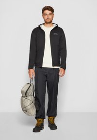 Giacca nera isolata, maglione beige e pantaloni neri. Cappuccio attaccato, chiusura zip e stivali neri con lacci gialli. Zaino grigio tenuto nella mano sinistra.
