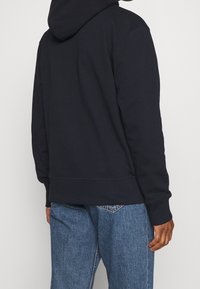 Tmavě modrá mikina s kapucí z měkké látky s volným střihem, vybavená kapsou klokaního typu, žebrovanými manžetami a neupraveným lemem. Kombinována s modrými džínovými kalhotami.