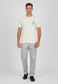 Krämfärgad t-shirt med en svart grafisk design på bröstet, parat med ljusgrå byxor och svarta skor, på en enfärgad bakgrund.