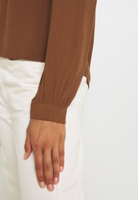 Main reposant sur le côté, portant un haut marron à manches longues avec poignets et un pantalon blanc devant un fond clair uni.