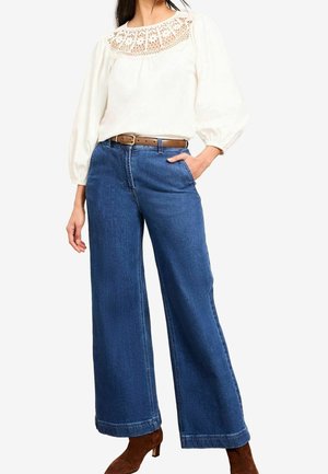 Femme portant un chemisier blanc avec un empiècement en dentelle, un jean bleu taille haute à jambes larges avec une ceinture marron, et des bottines marron.