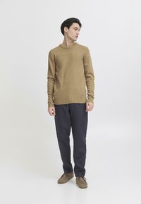 Lys brun genser med rund hals og ribbestrikkede mansjetter, kombinert med mørkeblå bukser og brune sko i et enkelt design.