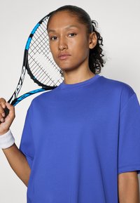 Lila sportpóló rövid ujjakkal és kerek nyakkivágással, amelyet egy személy visel, aki teniszütőt tart. A csuklóján izzadságszalag van.