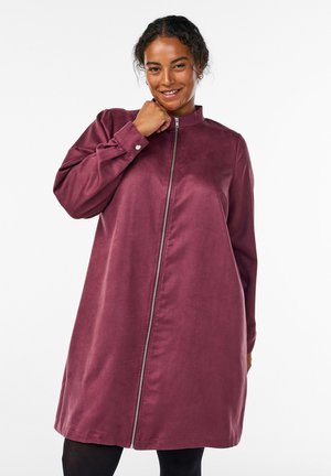 Donna che sorride, indossando un abito lungo con maniche, cerniera, di colore rosa scuro e colletto rialzato, su uno sfondo bianco semplice.