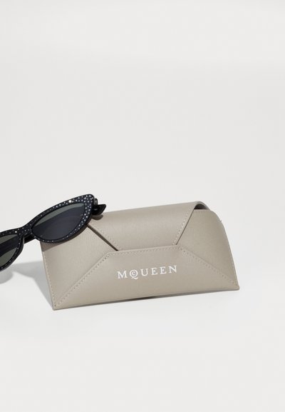 Etui de ochelari McQueen din piele bej, cu ochelari de soare negri în stil cat-eye, având rame împodobite cu strasuri, așezați deasupra.