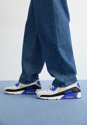 Person iført blå jeans og hvide sneakers med sorte, blå og beige detaljer, der står på en lyseblå gulv.