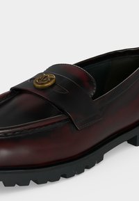 Mocassin en cuir marron foncé avec semelle crantée en caoutchouc noir et emblème en métal doré sur la bride avant, présenté en gros plan latéral incliné.