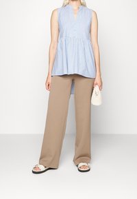 Tunique sans manches rayée bleu clair et blanche avec un col en V et des détails de boutons, associée à un large pantalon beige et des sandales beiges.