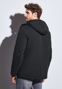 Černá zateplená bunda s kapucí, charakterizovaná hladkou texturou a minimalistickým designem. Zadní pohled ukazuje jemné detaily šití.