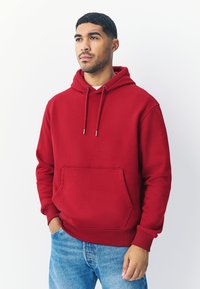 Rode hoodie gemaakt van zachte stof, met een voorzak, verstelbare capuchon met trekkoord en geribbelde manchetten. Gecombineerd met lichtblauwe spijkerbroek.