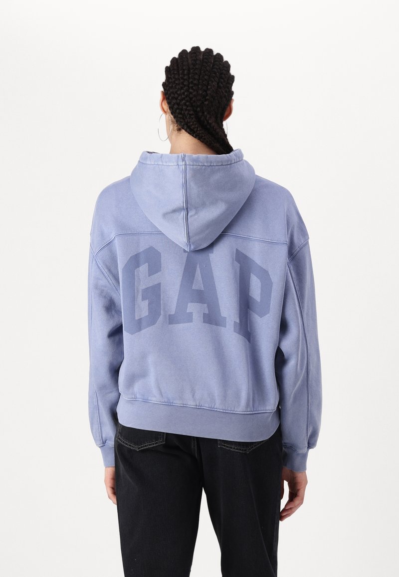 Personne aux cheveux tressés portant un sweat à capuche GAP violet clair et un pantalon noir, vue de dos sur un fond blanc uni.