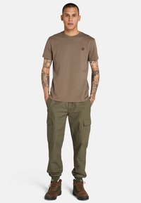 T-shirt in cotone marrone con scollo rotondo e logo. Pantaloni cargo verde oliva con tasche laterali. Scarponi da trekking marroni e verdi.