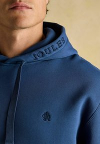 Sinine kapuutsiga pluus, millel on tikitud "Joules" kapuutsi peal ning väike logo rinna piirkonnas. Sujuv tekstuur ja vabaaja disain.