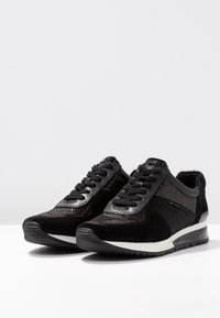 Black Michael Kors sneakers with textured and suede panels, black laces, and white-striped soles on a white surface.