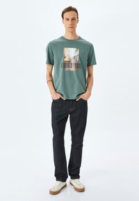 Camiseta de algodón verde con un estampado gráfico de una escena urbana y el texto "VISIÓN BORROSA". Combinada con jeans de denim oscuro y zapatillas.