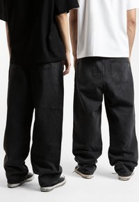 Black denim wide-leg pants with a relaxed fit, featuring back pockets and paired with black and white sneakers. Simple design, minimal detailing.