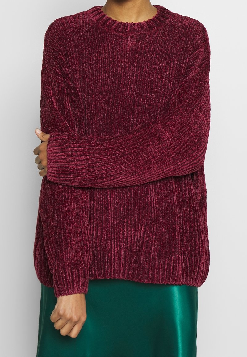 Vínový žebrovaný pletený svetr s dlouhými rukávy a kulatým výstřihem, s jemnou texturou a volným střihem. Doplněno zelenou sukní.