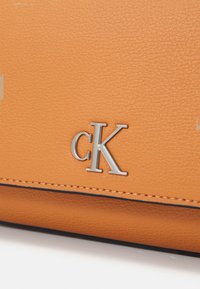 Borsa a tracolla in pelle arancione con texture, caratterizzata da un logo "cK" argentato e dettagli in cucitura nera. Vista ravvicinata con attenzione al materiale e al design.