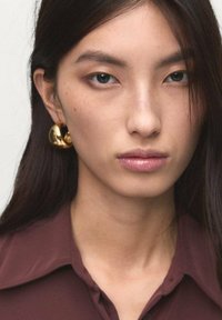 Primo piano del volto di una modella che indossa grandi orecchini a cerchio dorati. La modella ha i capelli scuri e lisci e indossa una camicia marrone con colletto.