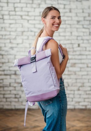 Eine lächelnde Frau mit einem weißen Oberteil und Jeans trägt einen helllila Rolltop-Rucksack über einer Schulter vor einer Backsteinmauer.