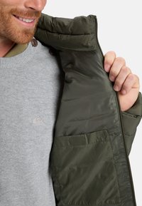 Hombre sonriendo, vistiendo un suéter gris claro con logo y sosteniendo una chaqueta acolchada verde oscura abierta, mostrando el bolsillo interior y la cremallera.